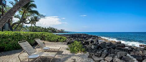 A beachside lounge with metal chairs and tables, a rocky shoreline, and palm trees.