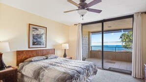 A hotel room with a bed, two bedside lamps, a ceiling fan, a framed picture on the wall, and a balcony with a view of the ocean.