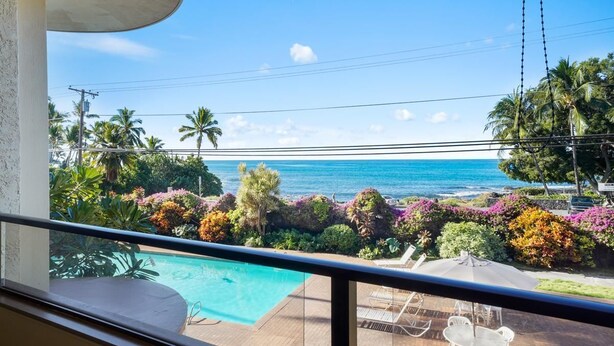 A balcony with a view of the ocean, a pool, and outdoor seating.