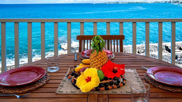 A wooden table set with plates, a glass, and a fruit arrangement on a balcony overlooking the ocean. - Sea Village 4-205 (Kailua-Kona)