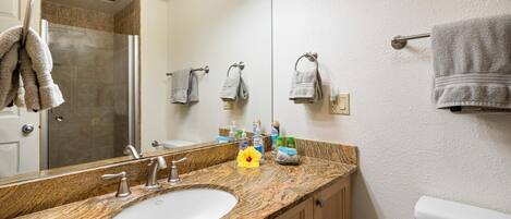 Un baño con encimera de granito, un lavabo blanco, un espejo y un inodoro. Hay toallas colgadas en un perchero y una ducha se ve al fondo.