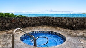 A stone-walled hot tub with a metal ladder, surrounded by a stone patio, overlooking a clear blue ocean. - Sea Village 4-205 (Kailua-Kona)