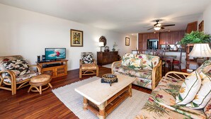 A living room with a television, wicker furniture, a dining area with a table and chairs, and a kitchen in the background. - Sea Village 4-205 (Kailua-Kona)
