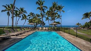 A rectangular pool with clear blue water, surrounded by lounge chairs and umbrellas, palm trees, and a view of the ocean. - Kona Isle C24 (Kailua-Kona)