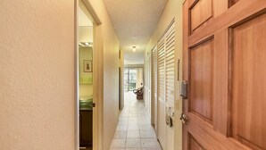 A hallway with a wooden door on the right, a bathroom with a toilet and sink on the left, and a balcony with a chair visible in the distance. - Kona Isle C24 (Kailua-Kona)