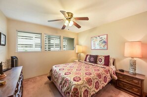 A bedroom with a bed, a ceiling fan, a nightstand with a lamp, a dresser, and a window with blinds.