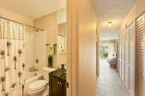 A bathroom with a shower, toilet, and sink, a hallway leading to a balcony, and a view of the outside.