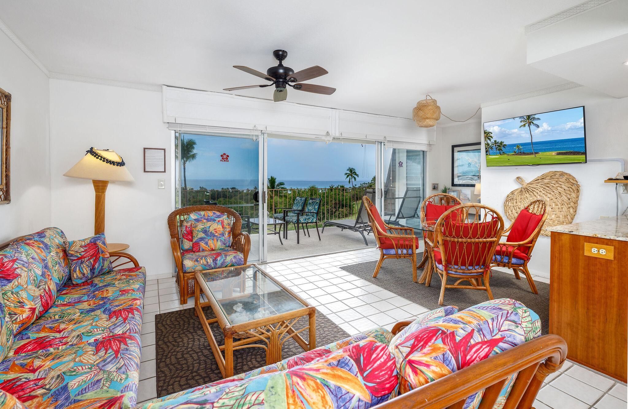 Amplia sala de estar con balcón, muebles de mimbre y un televisor mostrando una escena de playa tropical.