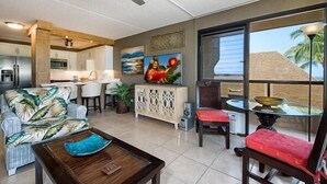 Un vaste espace de vie comprenant un canapé, une table basse et une table à manger avec des chaises. Au fond, on aperçoit un coin cuisine, une grande fenêtre donnant sur des palmiers et un ventilateur de plafond.