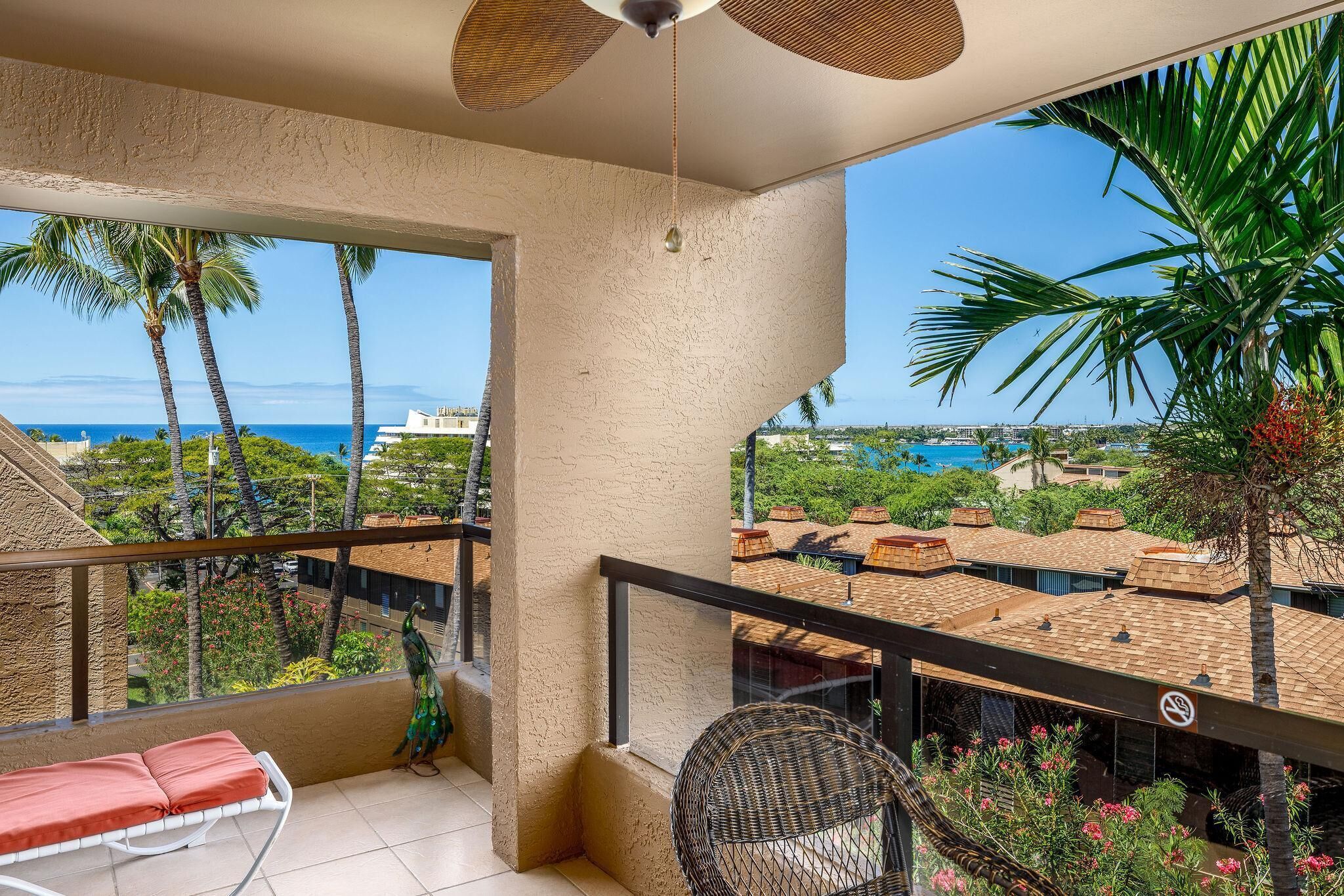 A balcony with a view of the ocean, palm trees, and a building with a tiled roof.