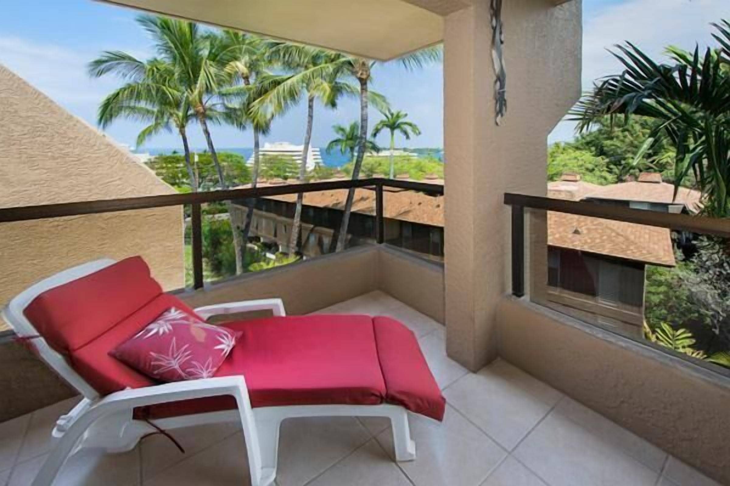 A balcony with a red lounge chair and a white chair, overlooking palm trees and a view of the ocean.