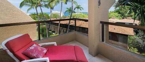 A balcony with a red lounge chair and a white chair, overlooking palm trees and a view of the ocean.