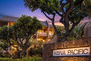 Een hotelschilt met de tekst 'KONA PACIFIC', omgeven door weelderige begroeiing en een gebouw met een modern ontwerp op de achtergrond.