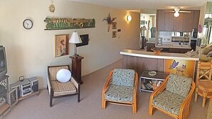 A living room with two armchairs, a sofa, a TV, and a kitchen area in the background. - Kalani Kai 105 (Kailua-Kona)