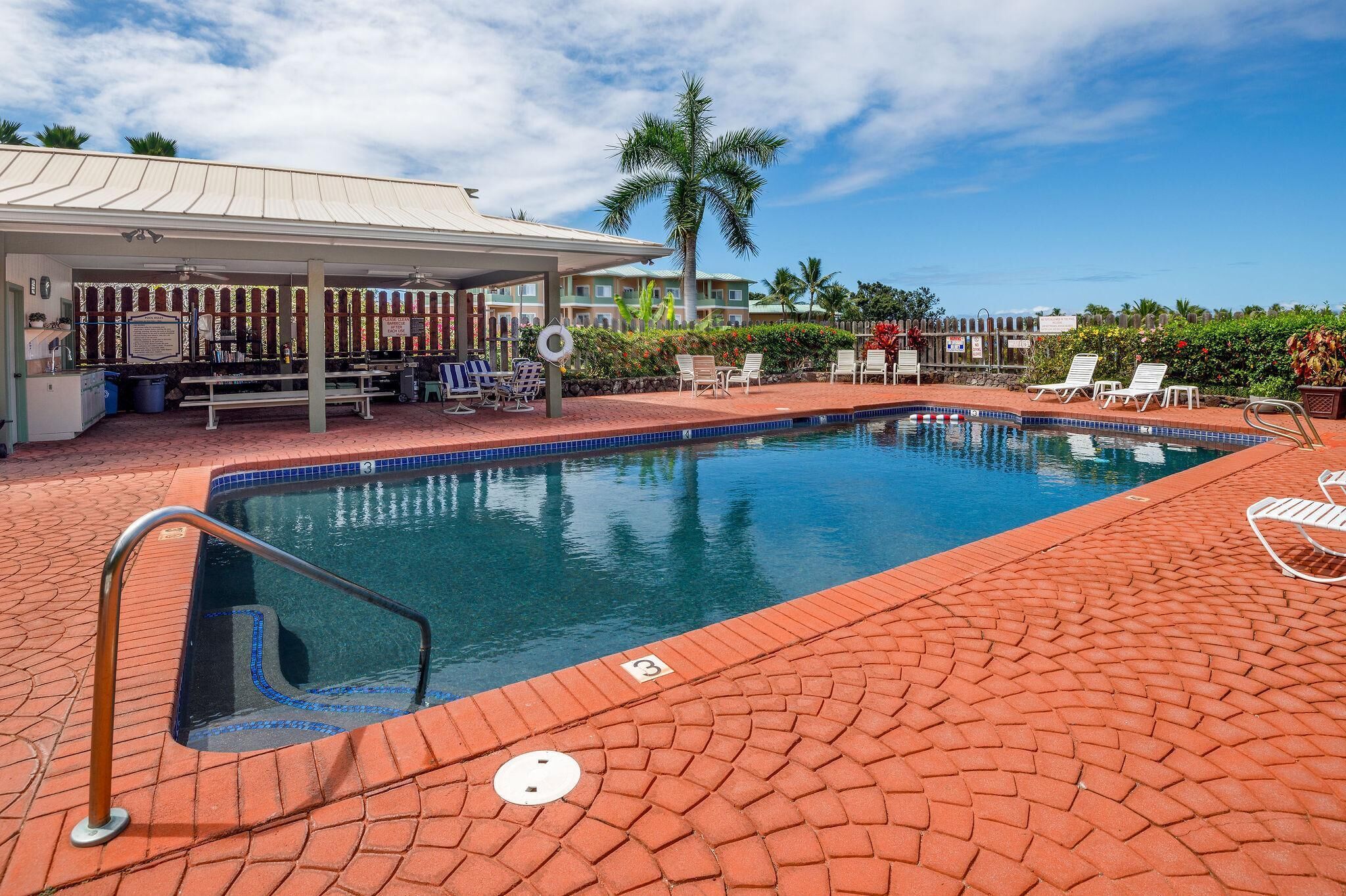 A swimming pool with a covered seating area, lounge chairs, and a palm tree in the background.