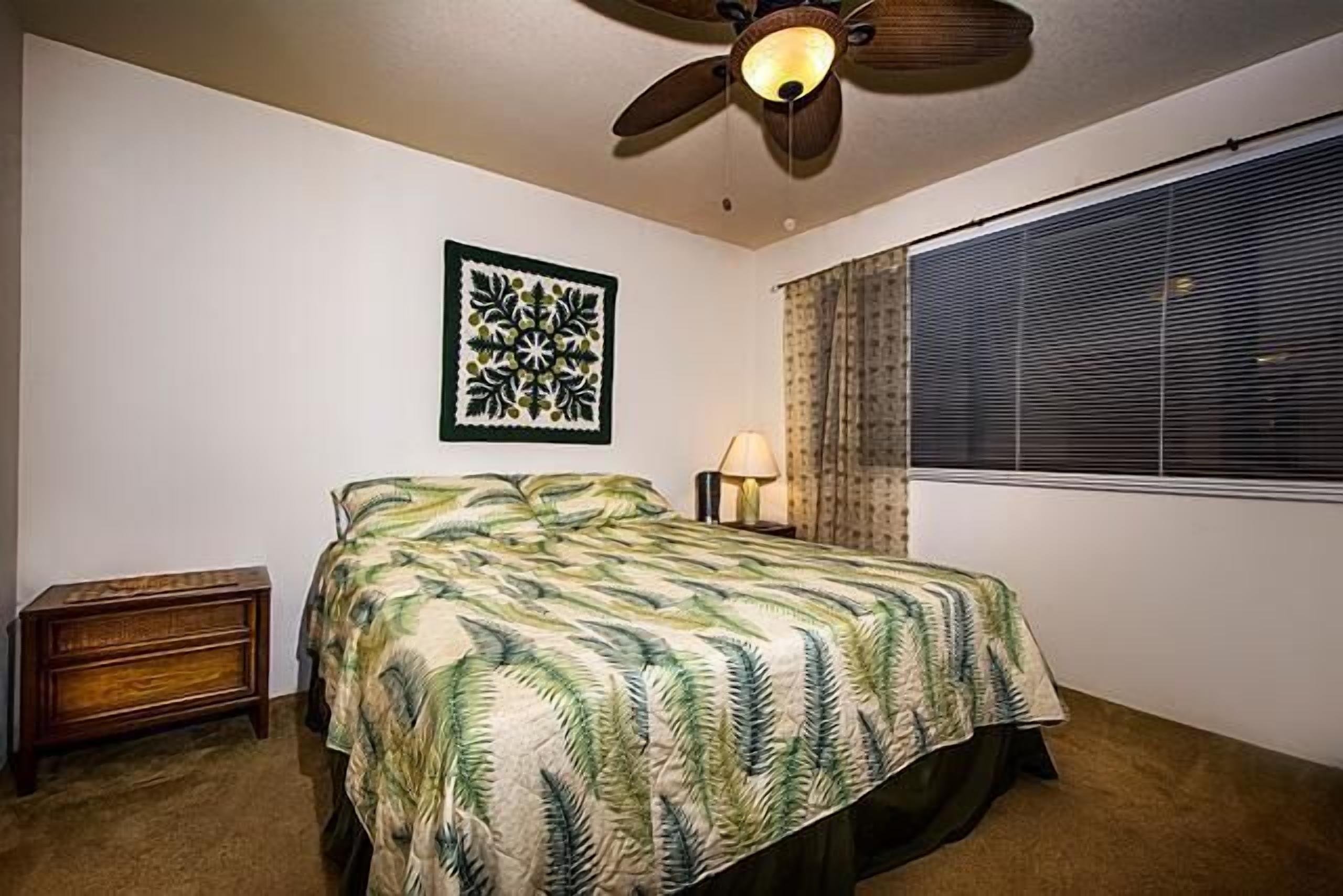 A bedroom with a bed, a ceiling fan, a window with blinds, a nightstand, and a wall art piece.
