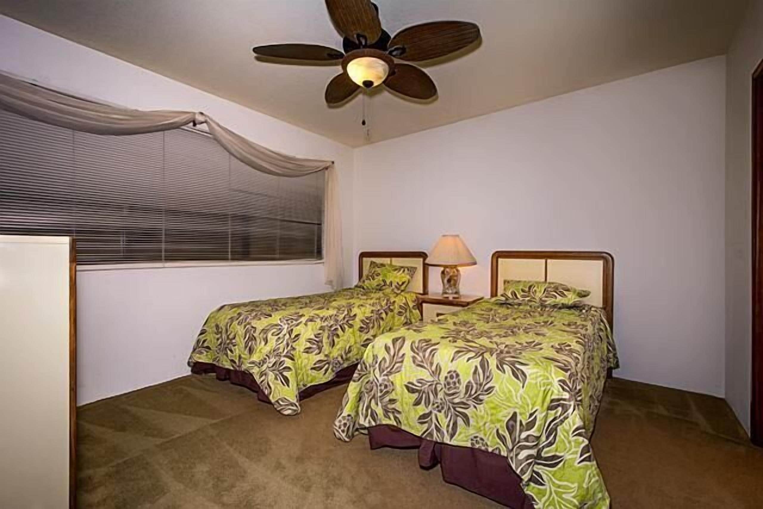 Makuuhuoneessa on kaksi sänkyä, kattotuuletin, ikkuna, jossa on sälekaihtimet, sekä yöpöytä, jossa on lamppu.