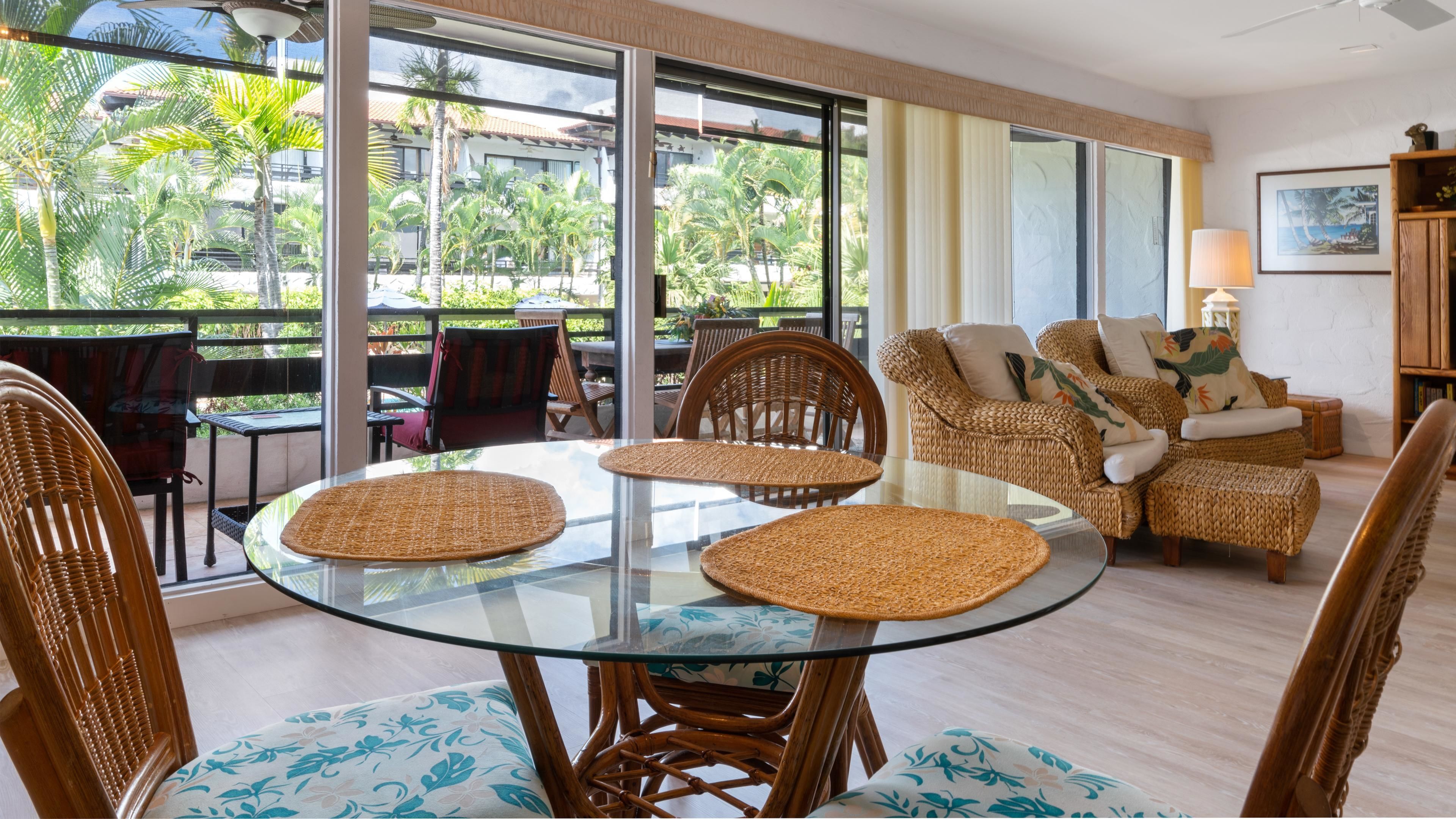 Ein modernes Wohnzimmer mit Glastisch, Rattanmöbeln und großen Fenstern mit Blick auf Palmen.