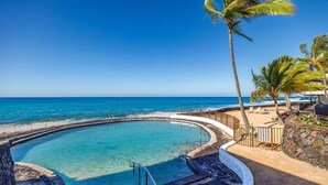 En rundlig infinitypool med kristallklart blått vatten, omgiven av en stenmur och palmer, med utsikt över en strand och havet.