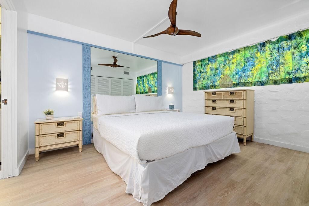 A bedroom with a large bed, a ceiling fan, a wooden dresser, and a wall with a floral tapestry.