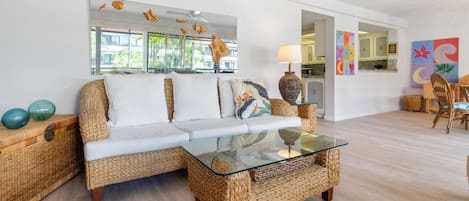 A living room with a wicker sofa, a glass coffee table, and a wicker side table. There is a painting on the wall and a view of the outside through large windows.
