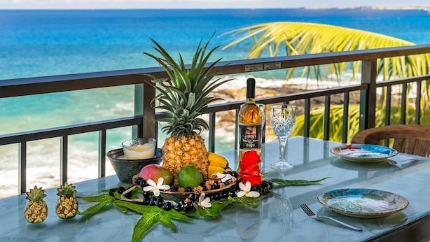Una mesa con frutas, una botella y un vaso en un balcón con vista al océano.