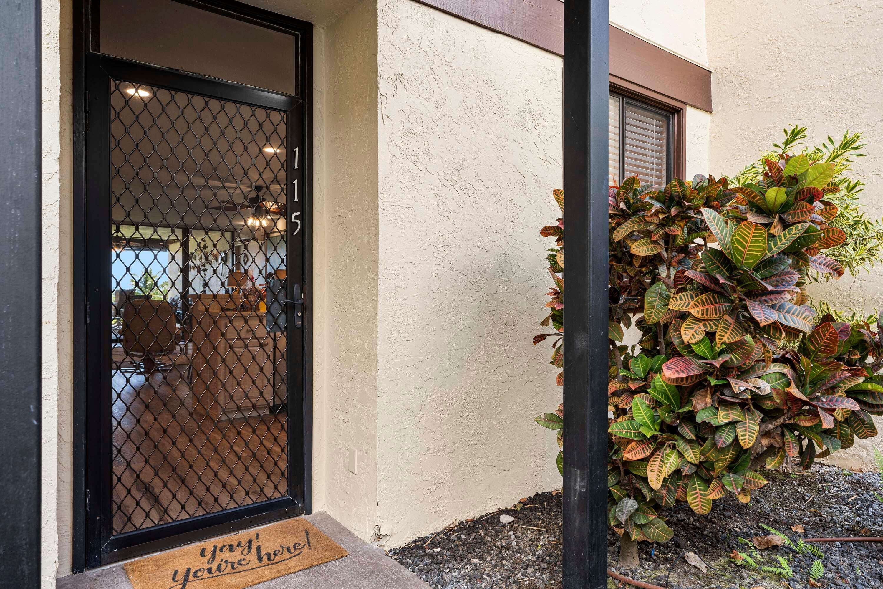 L’entrée d’un hôtel, avec une porte grillagée, un paillasson portant un message d’accueil et un pot de fleurs contenant une plante aux grandes feuilles colorées.