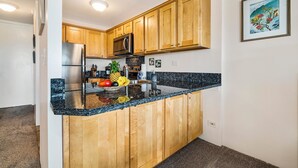 A kitchen with wooden cabinets, a stainless steel refrigerator, a microwave, and a granite countertop. - Kona Alii 608 (Kailua-Kona)