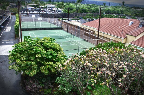 A tennis court surrounded by lush greenery and a parking area with vehicles.