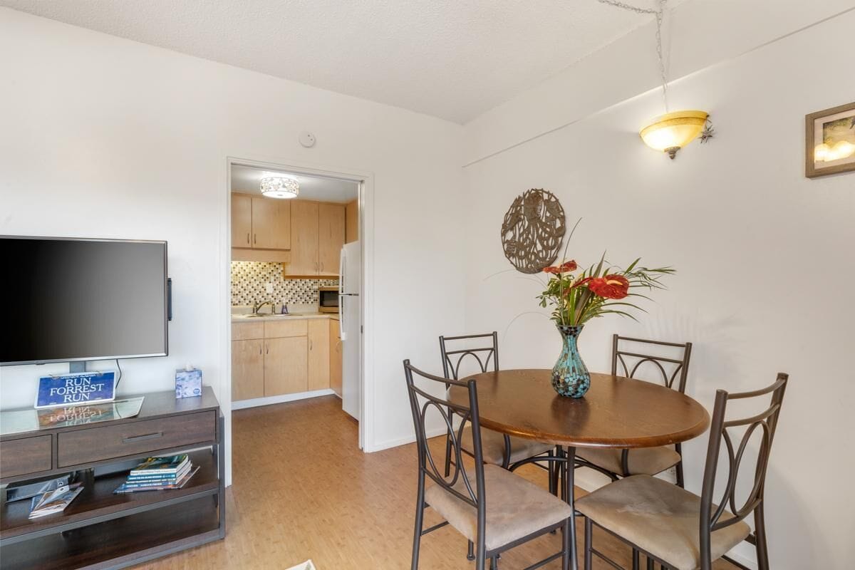 Un coin repas avec une table ronde, six chaises, un vase de fleurs, un téléviseur à écran plat posé sur un meuble, et une cuisine en arrière-plan.