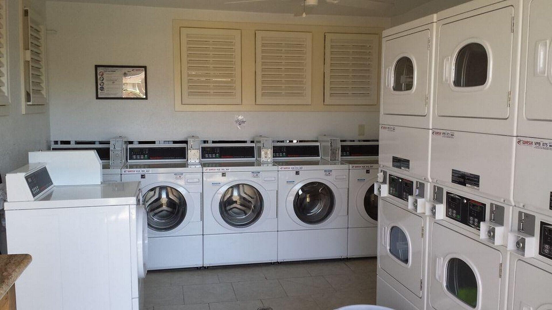 A laundry room with multiple washing machines and dryers, a control panel, and a window with blinds.