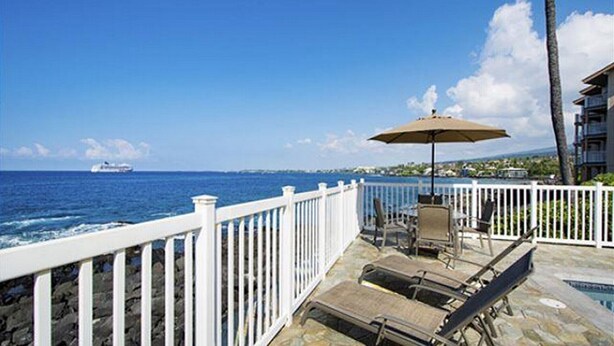 A deck with lounge chairs and an umbrella, overlooking a body of water with a ship in the distance. - Kona Shores#130 (Kailua-Kona)