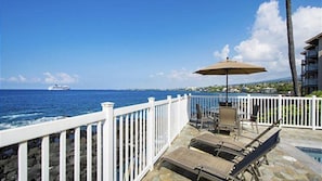 Une terrasse dotée de chaises longues et d’un parasol, offrant une vue sur une étendue d’eau où l’on aperçoit un navire au loin.