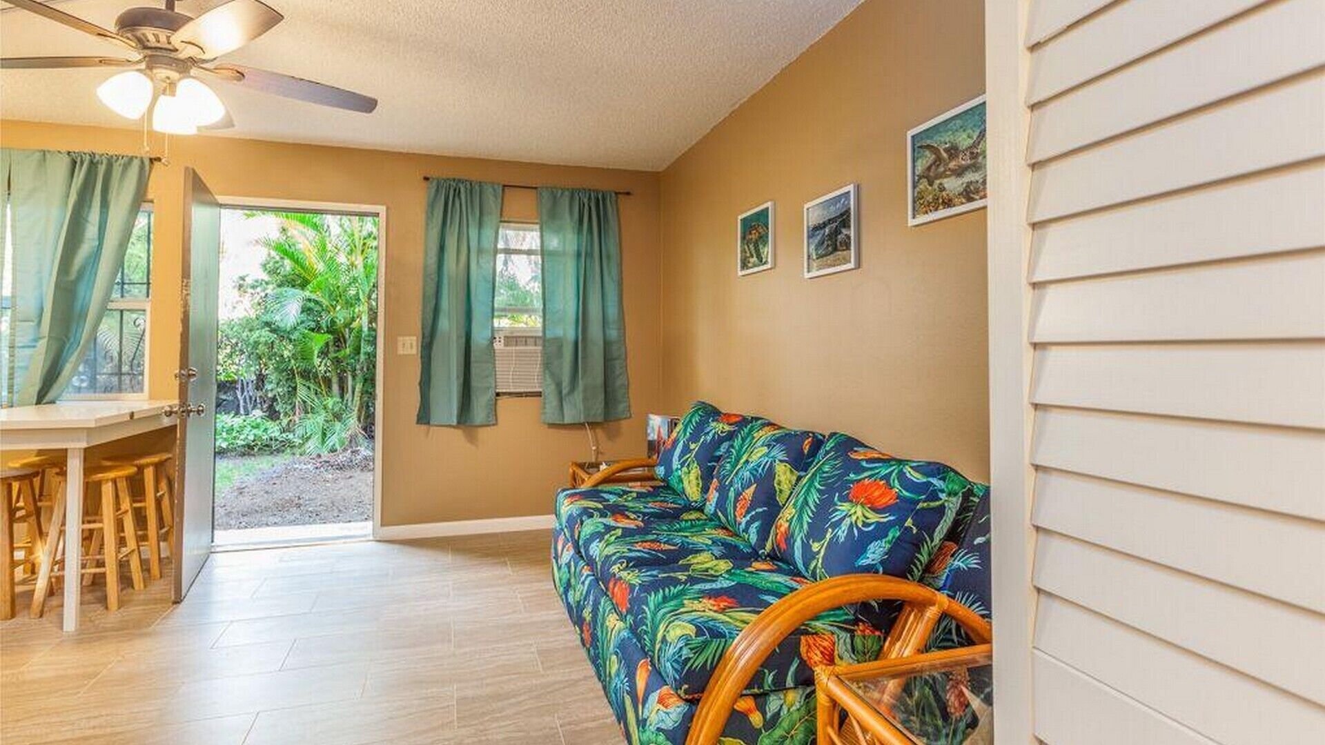 Ein Zimmer mit Holzboden, Deckenventilator, farbigem Sofa, Esstisch mit Stühlen und Blick ins Grüne.