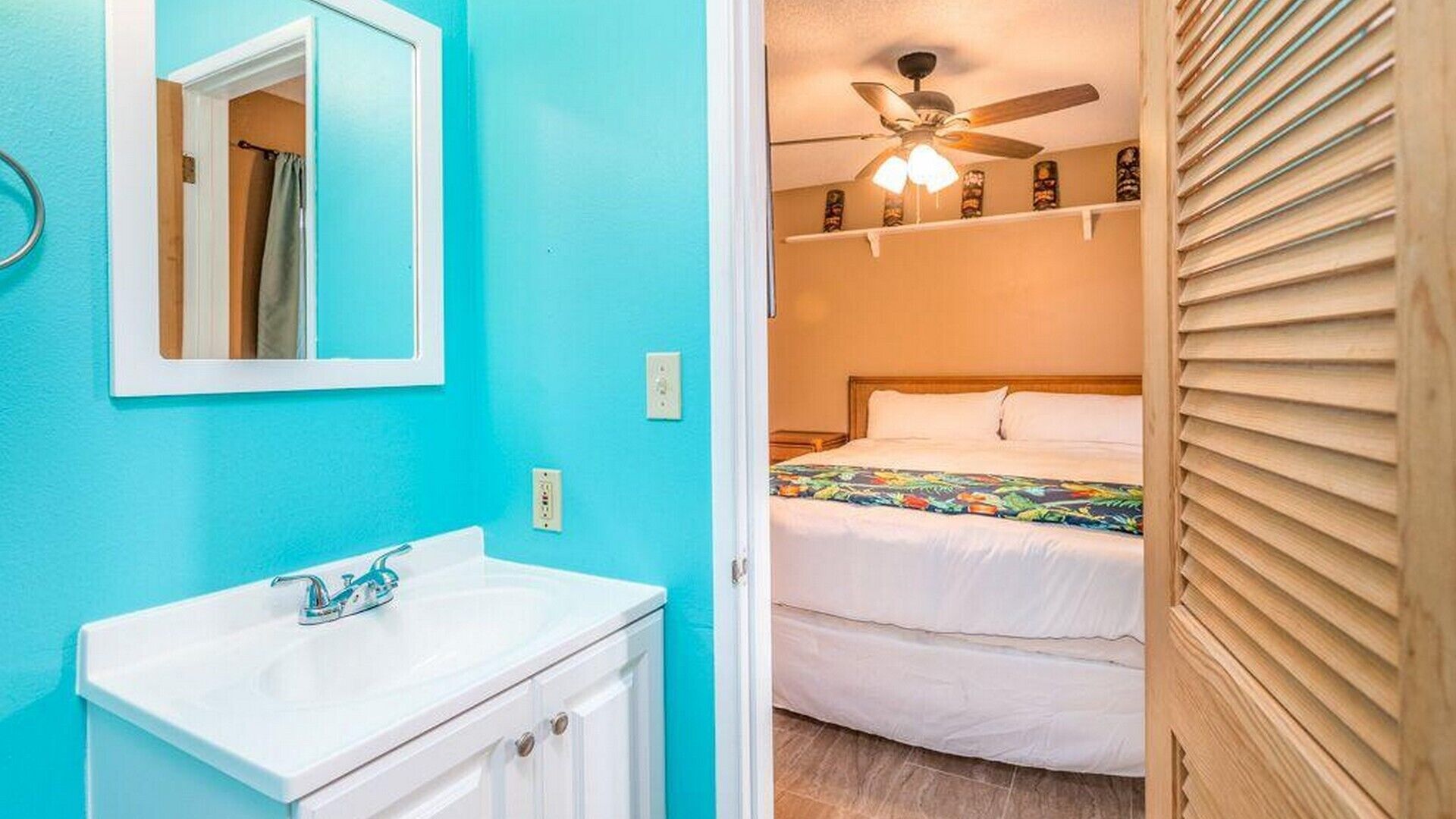 A room with a turquoise wall, a white sink, a mirror, a ceiling fan, and a bed with a floral bedspread.