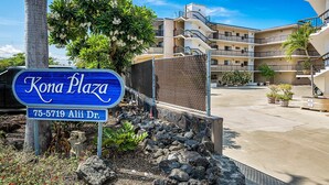 A sign for Kona Plaza with a multi-story building in the background.