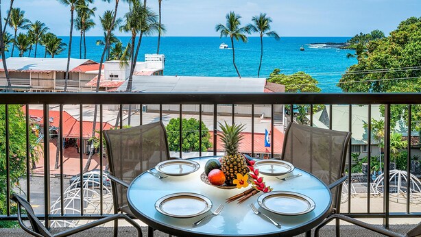 A balcony with a table set for four, overlooking a beach with palm trees and a boat in the distance.
