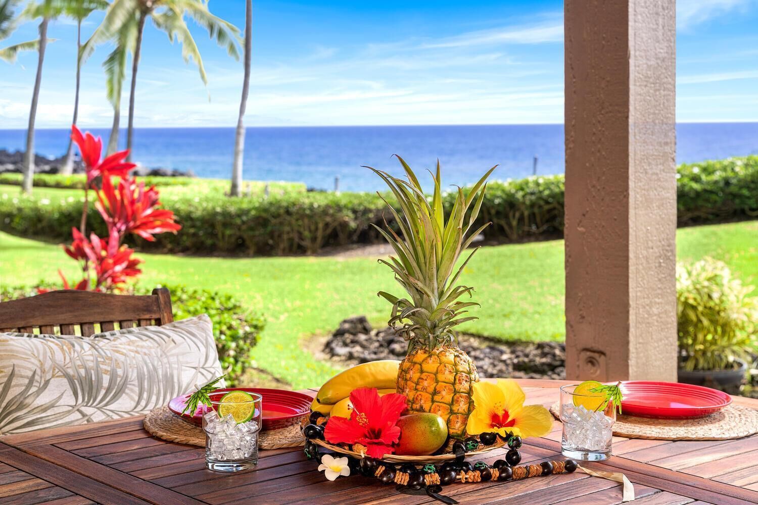 Una mesa de madera dispuesta con una piña, frutas, flores y bebidas, con vistas a un jardín exuberante y el océano.