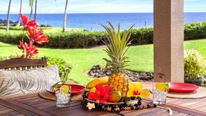 A wooden table set with a pineapple, fruits, flowers, and drinks, overlooking a lush garden and the ocean.