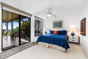 Une chambre à coucher comprenant un lit, un ventilateur de plafond, une table de chevet, une lampe, un tableau et une porte-fenêtre coulissante donnant sur un espace extérieur avec une chaise et une table.