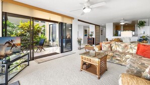 A spacious living room with a large glass door leading to a balcony, a sofa with tropical print, a coffee table, and a ceiling fan.