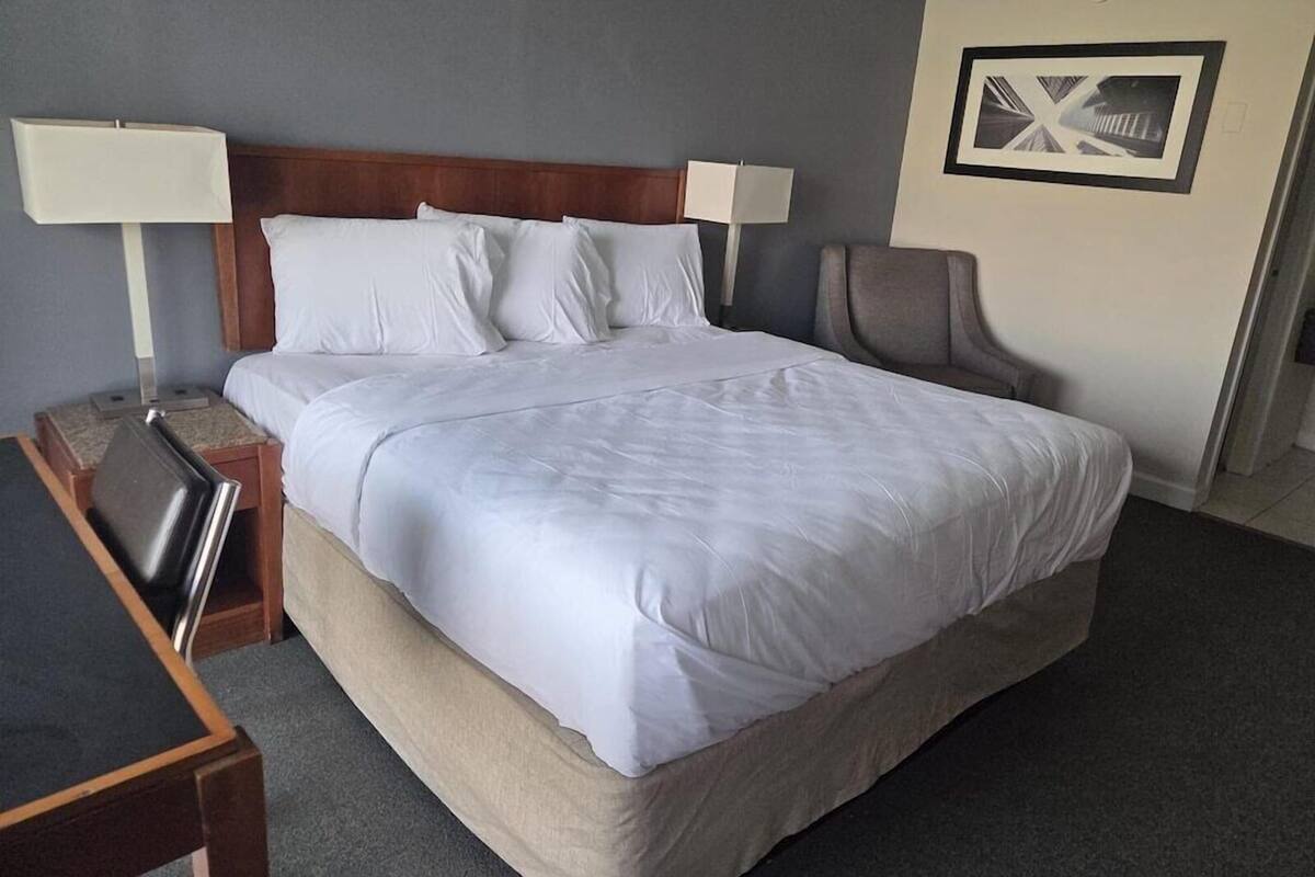 A hotel room with a large bed, two bedside lamps, a chair, a desk, and a framed picture on the wall.