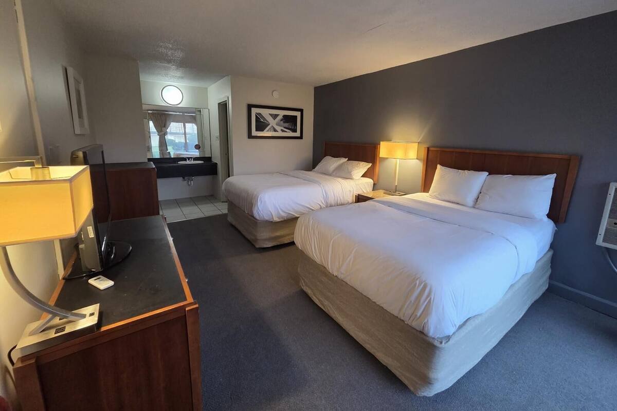 A hotel room with two beds, a desk, a chair, a sink, and a mirror.
