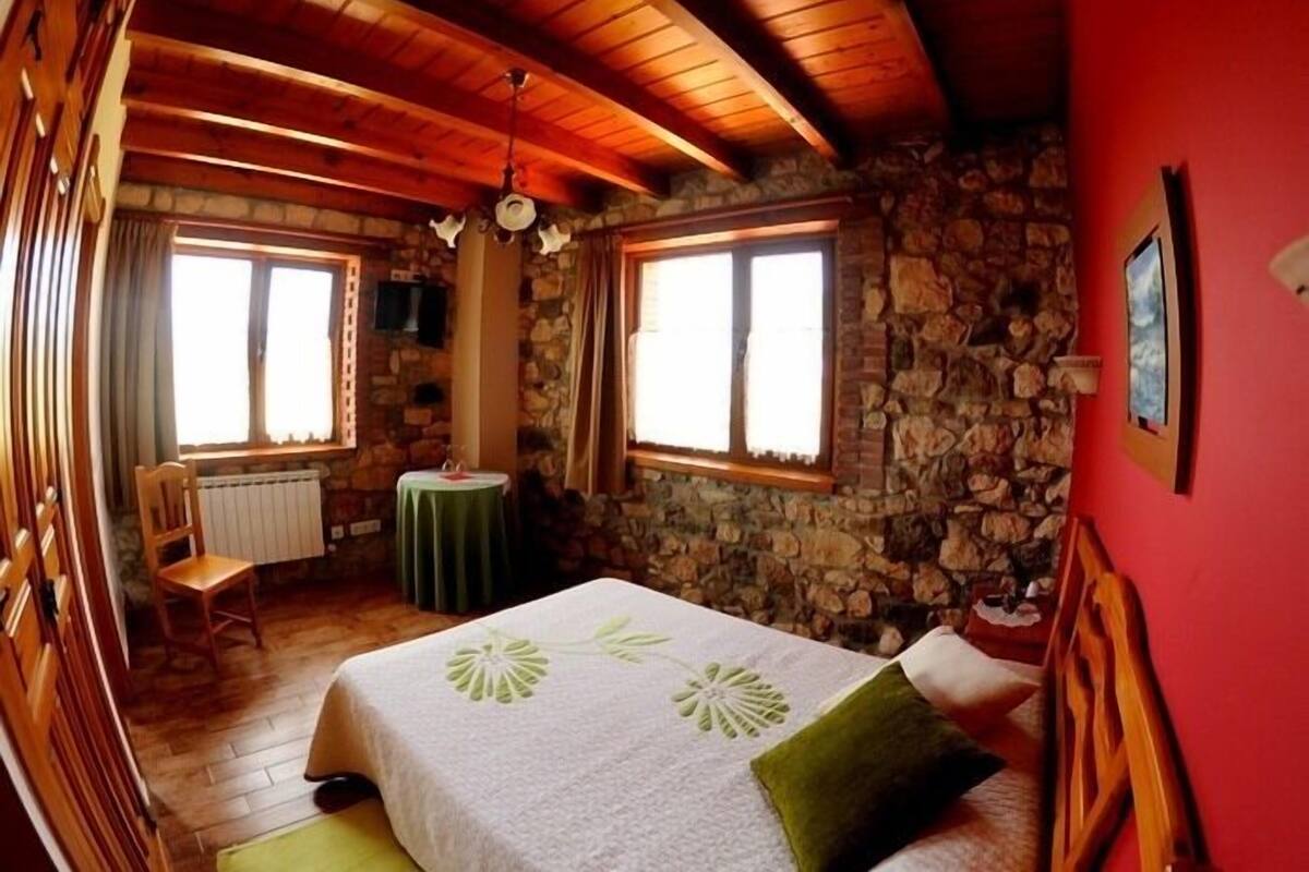 A cozy room with a stone wall, wooden ceiling beams, a bed with a green pillow, a small table, and a chair.