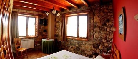 A cozy room with a stone wall, wooden ceiling beams, a bed with a green pillow, a small table, and a chair.