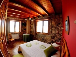 Un cuarto acogedor con pared de piedra, vigas de madera en el techo, una cama con almohada verde, una mesita y una silla.