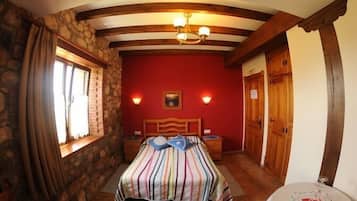 Un dormitori amb paret de pedra, bigues de fusta, un llit amb estriatges, dues taules de nit, una petita taula auxiliar, una porta de fusta i una finestra amb cortines.