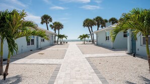 Ein Pfad führt an Reetdachbungalows am Strand entlang, mit Palmen und freiem Blick auf den Ozean.