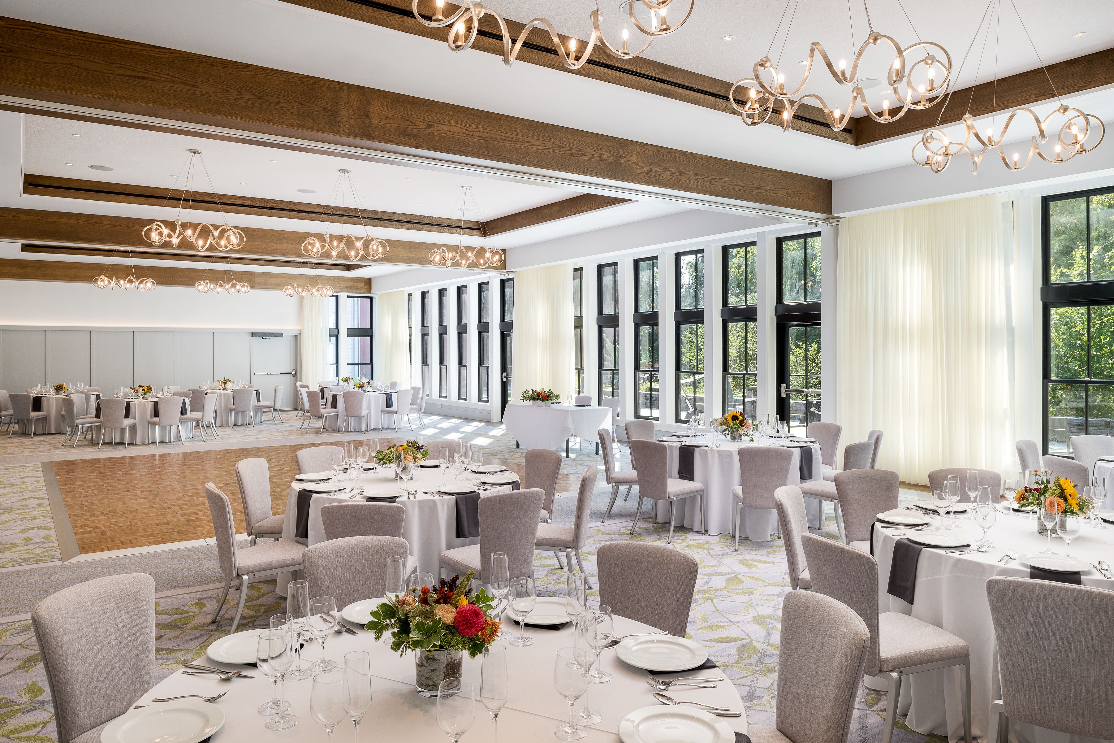 A spacious banquet hall with round tables set for dining, large windows, and modern chandeliers.
