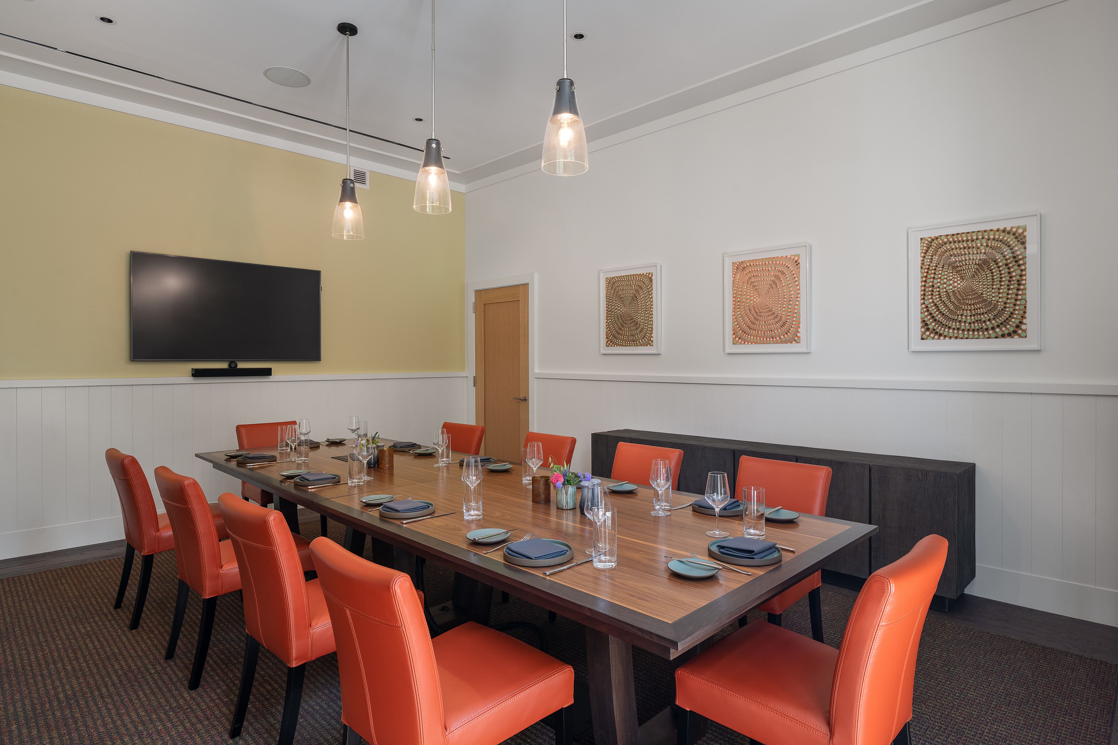 A conference room with a long table, orange chairs, a flat-screen TV, pendant lights, and woven wall art.